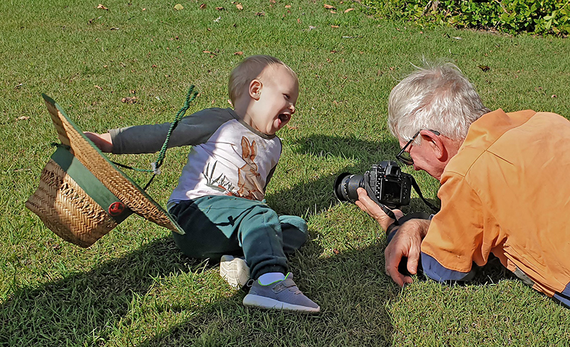 Kay Suter 2nd place photograph. A small child sits on the grass, laughing and holding a straw hat, while their grandparent takes their photograph.
