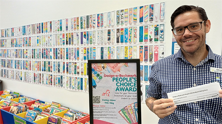 A library staff member stands in front of a wall filled with competition entries.