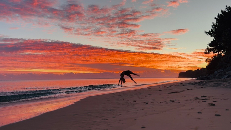 Photo taken by Cailee Steen, titled Dancing with the Tide. Person doing a flip on the sandy beach at dawn. 