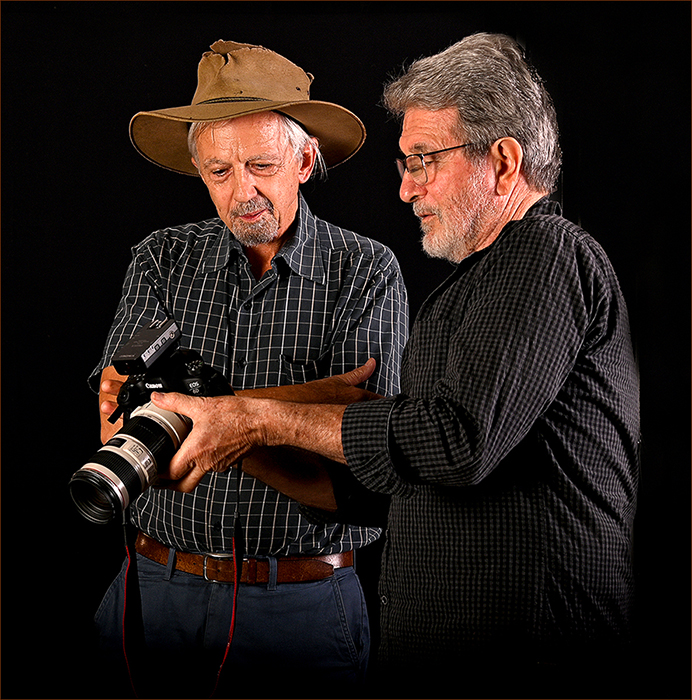 Greg Suter 1st place photograph. Two men standing in front of a dark backdrop looking at the screen of a digital camera. 