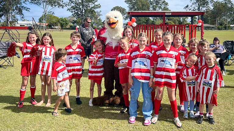 A large group of young people from the Tinana Football Club, dressed in red and white uniforms, pose with their mascot.